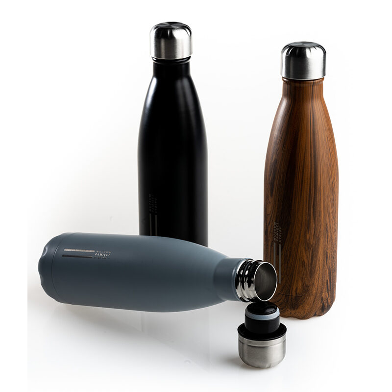 Zdjęcie: Na białym tle trzy butelki termiczne. Butelki są w różnych wariantach kolorystycznych: czarnym, drewnianym i szarym. Zakrętki butelek są w kolorze metalicznym. Na pierwszym planie szara butelka. Jest otwarta. Leży oparta o zakrętkę. Na drugim planie obok siebie butelka czarna i w kolorze drewna.