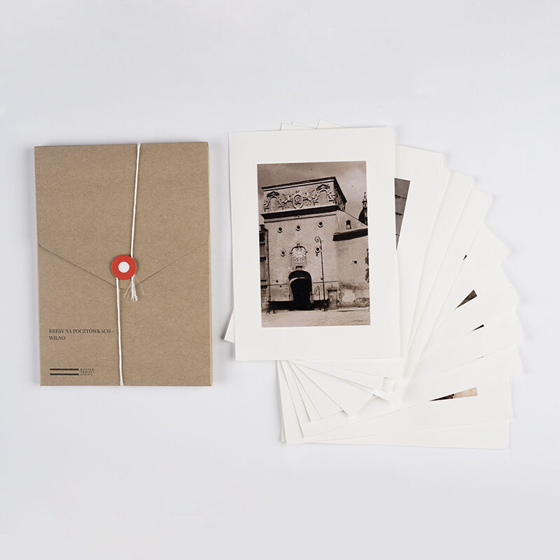 Zdjęcie: Na białym tle brązowa teka. Obok niej pocztówki. Rozłożone pocztówki tworzą wachlarz. Najlepiej widoczna jest pierwsza pocztówka przedstawiająca Ostrą bramę w Wilnie.