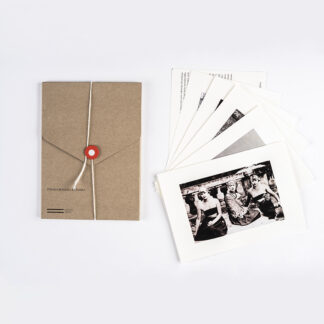 Zdjęcie: Na białym tle brązowa teka. Obok niej pocztówki. Rozłożone pocztówki tworzą wachlarz. Najlepiej widoczna jest pierwsza pocztówka przedstawiająca trzy kobiety siedzące na podłodze tarasu.